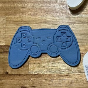 Peut inclure: Deux emportes-pièces en plastique blanc en forme de manette de jeu vidéo. Un emporte-pièce est plein et l'autre est un contour avec une petite image de manette à l'intérieur. Une manette en plastique bleue est également représentée.