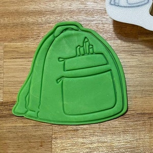 May include: A green plastic cookie cutter shaped like a backpack with a zipper and a pocket. The cutter is on a wooden surface next to a white plastic cookie cutter shaped like a backpack. The white cutter has the text "Backpack | GBBB8" on it.