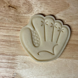 Peut inclure: Emporte-pièce beige en forme de gant de baseball sur une surface en bois. Le découpoir a la forme d'un gant de baseball, avec des lignes détaillées pour montrer les doigts et la toile. Le fond est une surface en bois brun clair.