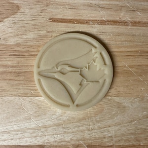 May include: A circular, beige cookie cutter featuring a bird's head and a maple leaf design within a border. The cutter sits on a light brown wooden surface.