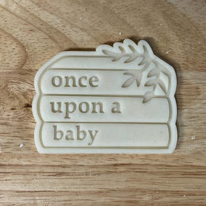 Peut inclure: Emporte-pièce beige en forme de pile de livres avec les mots "once upon a baby" et un motif de feuilles. L'emporte-pièce est posé sur une surface en bois.