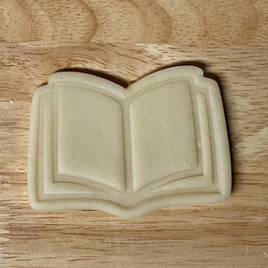May include: A light-coloured, open book-shaped cookie cutter. The cutter is made of a light-coloured material and has a detailed outline of an open book with visible pages. The cutter is designed for baking biscuits.