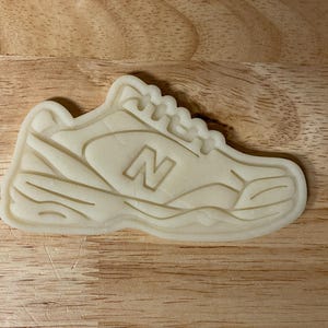 以下が含まれることがあります： スニーカーの形をした、明るいベージュ色のクッキーカッター。スニーカーの側面には目立つ「N」のロゴがあり、靴のデザインを表す詳細な線があります。クッキーカッターは木製の表面にあります。