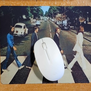May include: A white computer mouse with a black scroll wheel and a black button on the top. The mouse is on a black and white mousepad with a picture of the Beatles crossing a street.