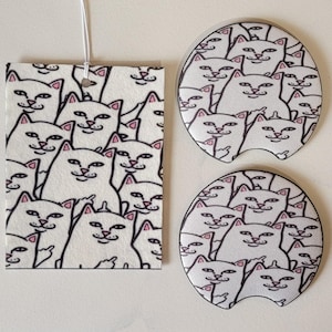 May include: A white air freshener and two car coasters with a repeating pattern of white cats with pink ears and noses. The cats are giving the middle finger. The air freshener is rectangular and the coasters are circular.