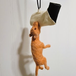 May include: A small, orange, felt dog toy hanging from a white and black cord. The dog is suspended upside down with its legs in the air.