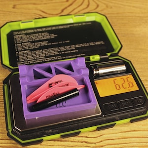 Pode incluir: Uma balança digital preta com uma caixa verde, aberta para revelar uma bandeja roxa com três penas rosa e um eixo de flecha preto. A balança marca 6,26.