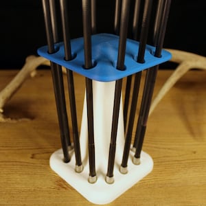 Peut inclure: Un support de flèches blanc et bleu avec des flèches noires. Le support a une base triangulaire et un dessus bleu avec des trous pour les flèches. Les flèches sont noires avec des pointes argentées. L'arrière-plan comprend une surface en bois et un bois de cerf.