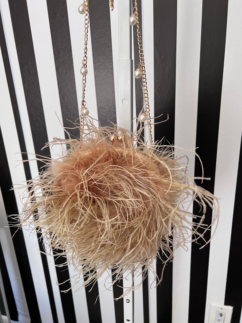 May include: A small, round, beige clutch bag with a fluffy, ostrich feather exterior. The bag has a gold chain strap with pearl accents.