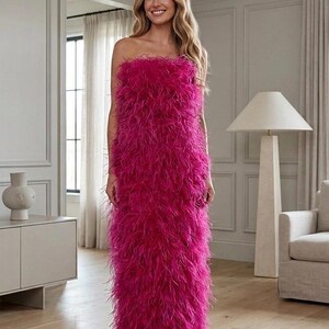 May include: A strapless, floor-length fuchsia dress covered in soft, fluffy feathers. The dress has a straight silhouette and is the focal point of the image. The background features a neutral-toned room with a modern aesthetic.