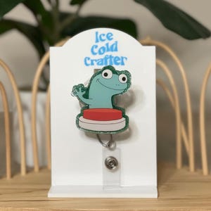 Puede incluir: Un portatarjetas con un diseño de rana de dibujos animados. La rana es de color azul claro con ojos grandes y está de pie sobre un botón rojo. El texto "Ice Cold Crafter" está encima de la rana. El carrete está unido a una tarjeta blanca.