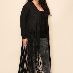 May include: A long black fringe cardigan. The cardigan features long fringe detailing along the front and bottom hem. The model is wearing a black top and grey patterned trousers. The cardigan is open at the front.