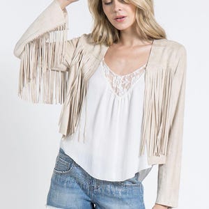 Könnte beinhalten: Beige Jacke in Wildlederoptik mit langen Fransen an den Ärmeln und der Vorderseite. Die Jacke wird über einem weißen Oberteil getragen und mit Jeansshorts kombiniert. Die Shorts haben einen ausgefransten Saum und sind mit kleinen, glänzenden Details verziert.