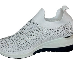 May include: White slip-on sneaker with a wedge heel. The shoe is covered in small, sparkling rhinestones. The sole is white with a black bottom and a silver accent on the heel.