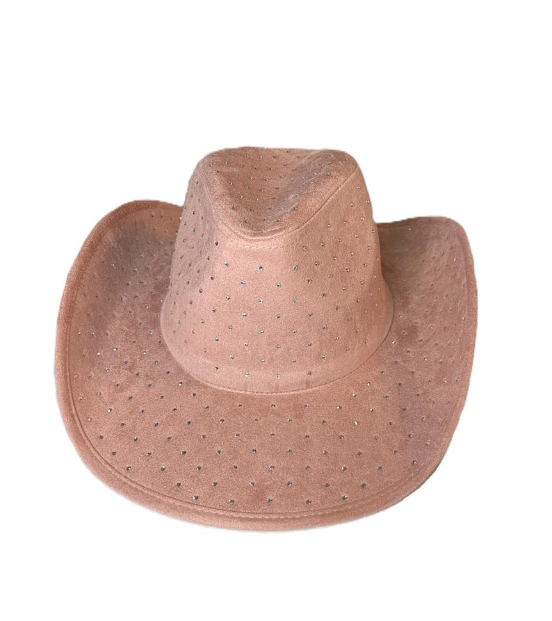 May include: A blush-colored cowboy hat with a suede-like texture. The hat is adorned with small, evenly spaced rhinestones across the crown and brim. The hat has a classic cowboy hat shape with a rounded crown and a wide, curved brim.