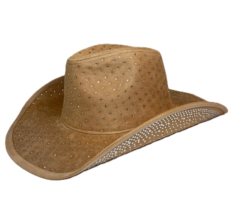 May include: A tan cowboy hat with a wide brim and a rounded crown. The hat is adorned with small, sparkling rhinestones scattered across the surface and along the brim. The hat is made of a suede-like material.