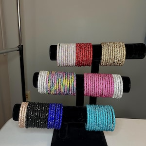 Puede incluir: Un expositor negro exhibe múltiples pilas de pulseras coloridas. Las pulseras son de varios colores, incluyendo plata, rojo, oro, arcoíris, rosa, negro, azul y turquesa. Las pulseras se exhiben en un expositor de terciopelo negro.