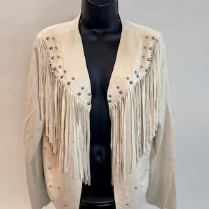 Puede incluir: Chaqueta de ante beige con flecos en el pecho y los hombros. La chaqueta presenta tachuelas plateadas a lo largo de los flecos y el cuello. La chaqueta se muestra en un maniquí negro.