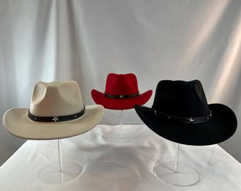 Puede incluir: Tres sombreros de vaquero en crema, rojo y negro, cada uno con una banda negra adornada con estrellas plateadas. Los sombreros se exhiben en soportes acrílicos transparentes, mostrando su clásico estilo western.