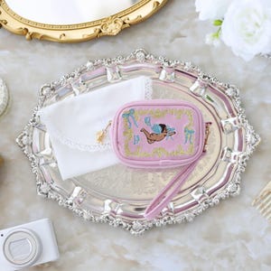 May include: A pink, rectangular coin purse with an embroidered cherub design, gold trim, and a wrist strap. It rests on a silver tray with a white lace-trimmed pouch. A gold-framed mirror and a digital camera are also in the image.