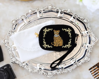 May include: A black velvet clutch with a gold embroidered leopard design and a wrist strap sits on a silver tray. A white lace-trimmed handkerchief and a gold-framed mirror are also visible.