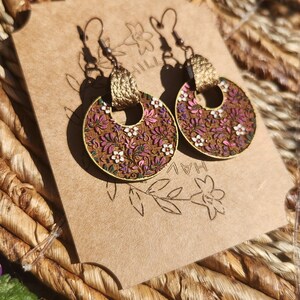 May include: A pair of gold hoop earrings with a floral pattern. The earrings are made of wood and have a brown background with pink, white, and green flowers. The earrings are hanging on a brown card with the text "HAL" and "HILL" visible.