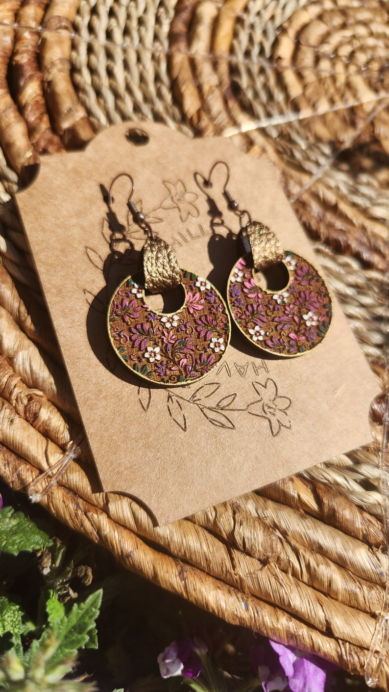 May include: A pair of gold hoop earrings with a floral pattern. The earrings are made of wood and have a brown background with pink, white, and green flowers. The earrings are hanging on a brown card with the text "HAL" and "HILL" visible.