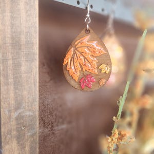 May include: Wooden teardrop earrings with a fall leaf design. The earrings feature a large orange leaf with smaller red and yellow leaves. The earrings are hanging from a decorative metal display.