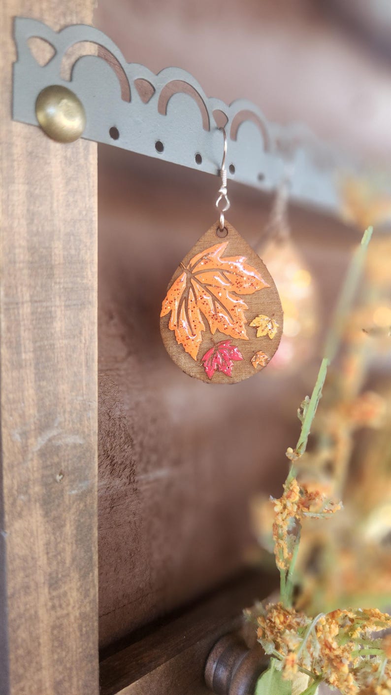 May include: Wooden teardrop earrings with a fall leaf design. The earrings feature a large orange leaf with smaller red and yellow leaves. The earrings are hanging from a decorative metal display.