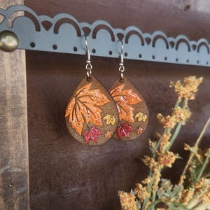 May include: Wooden teardrop earrings with a fall leaf design. The earrings feature orange, red, and yellow leaves with glitter accents. The earrings hang from silver-colored hooks, perfect for autumn.