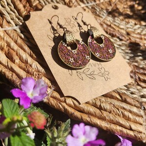 May include: A pair of round, floral earrings with a brown background and pink, white, and green flowers. The earrings have a gold-colored metal hoop and a brown tassel hanging from the hoop. The earrings are displayed on a brown paper card with the word "HAV" written on it.