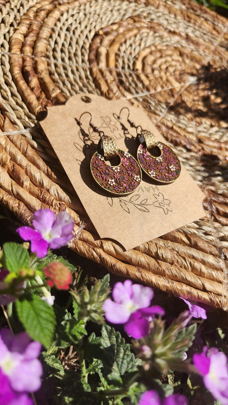 May include: A pair of round, floral earrings with a brown background and pink, white, and green flowers. The earrings have a gold-colored metal hoop and a brown tassel hanging from the hoop. The earrings are displayed on a brown paper card with the word "HAV" written on it.