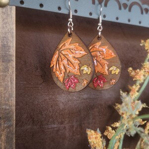 May include: Wooden teardrop earrings with a fall leaf design. The earrings feature a large orange leaf with smaller red and yellow leaves. The earrings are hanging from silver hooks.