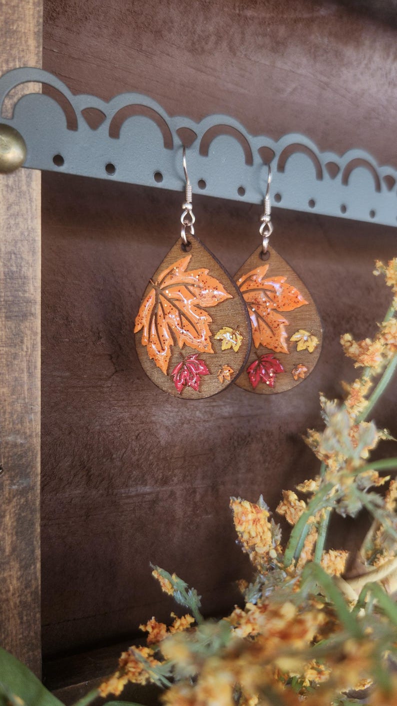 May include: Wooden teardrop earrings with a fall leaf design. The earrings feature a large orange leaf with smaller red and yellow leaves. The earrings are hanging from silver hooks.