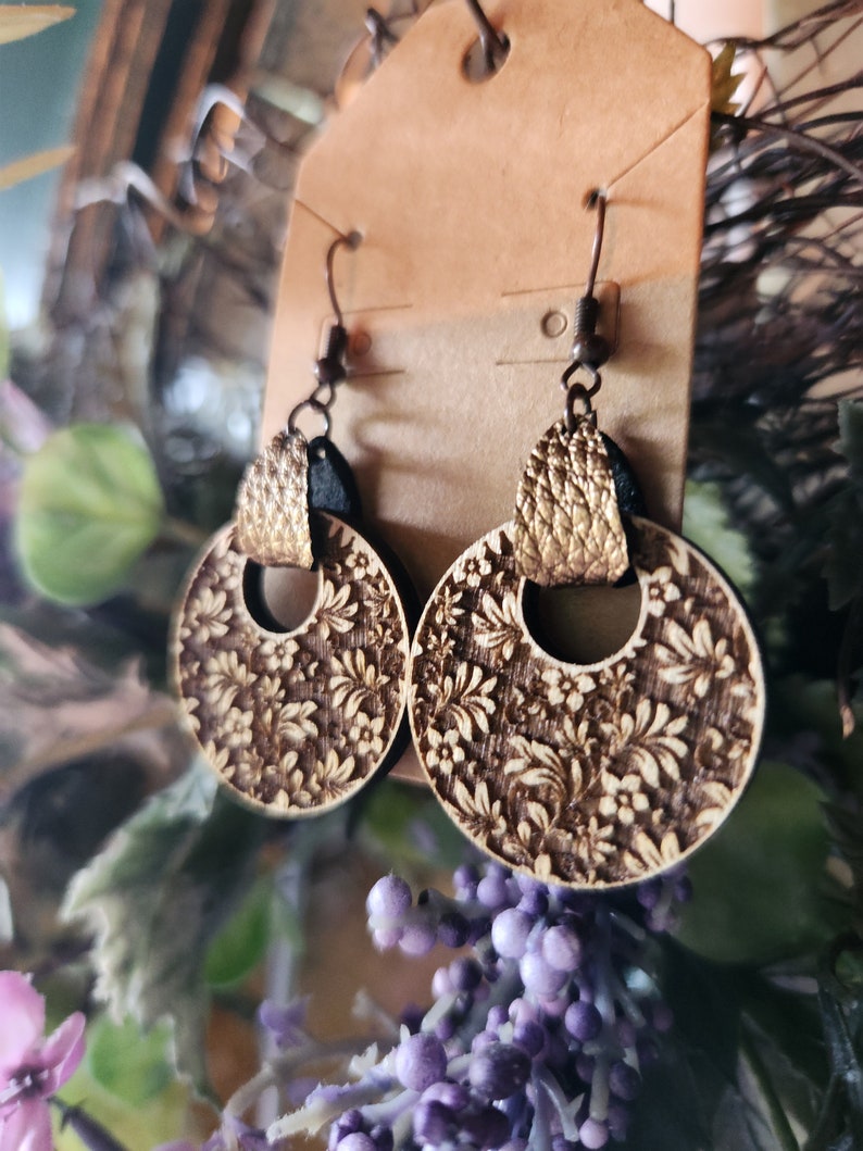 May include: A pair of wooden earrings with a floral design. The earrings are brown with gold accents and have a circular shape.