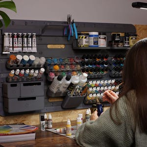Puede incluir: Un sistema organizador modular de pared en color gris oscuro para manualidades, con múltiples estantes, soportes para botes de pintura, cajones y un portaherramientas. El sistema contiene varios tubos de pintura, frascos y pinceles.