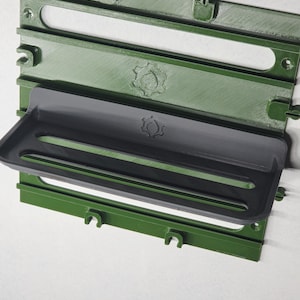 May include: A dark green wall-mounted storage panel featuring horizontal slots and integrated hooks, with a detachable dark gray shelf unit attached to the center. The panel and shelf both display a small gear-shaped logo embossed on their surfaces.