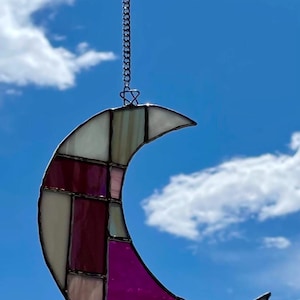 May include: Stained glass crescent moon suncatcher with a chain for hanging. The moon is composed of various shades of pink, white, and purple glass pieces. The background is a bright blue sky with fluffy white clouds.