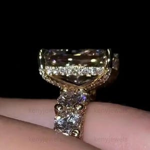 Può includere: Primo piano di un anello d'oro con una grande gemma rettangolare di colore chiaro. L'anello presenta un design dettagliato e ornato con pietre più piccole attorno alla gemma principale e sulla fascia. Lo sfondo è scuro.