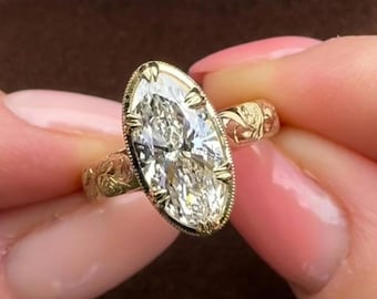 Könnte beinhalten: Ein Goldring mit einem großen, im Marquise-Schliff facettierten Mittelstein, der von Krappen gehalten wird. Die Ringschiene besteht aus Gelbgold und zeigt filigrane, eingravierte Blumenmuster an den Seiten sowie einen Milgrain-Rand.