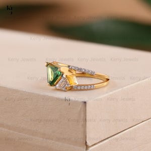 A double-band yellow gold ring holds a large, triangular green stone in a three-prong setting. A small cluster of sparkling white diamonds is set on the lower band right next to the central green gem.