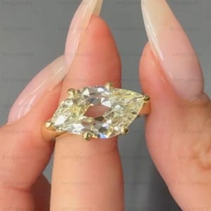 May include: A gold ring featuring a large, elongated marquise-cut gemstone with a faceted surface. The stone is held in place by multiple small prongs on a simple, polished gold band. The design highlights the clear, reflective quality of the stone.