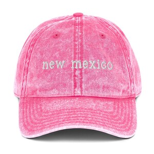 May include: Pink distressed denim baseball cap with white embroidered text that reads "new mexico".