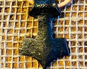 May include: A black metal pendant in the shape of a hammer, hanging from a brown leather cord. The pendant is a symbol of Thor, the Norse god of thunder.