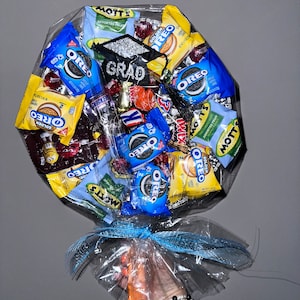 May include: A graduation gift bouquet wrapped in clear plastic with a blue ribbon. The bouquet is filled with various snacks, including Oreo cookies, Mott's fruit snacks, and candy. A black graduation cap with the word "GRAD" is on top of the bouquet.