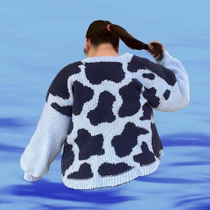 Puede incluir: Un suéter de punto con un estampado de vaca. El suéter presenta una base azul claro con manchas azul oscuro. Las mangas son de color azul claro liso. La persona de la imagen lleva el suéter.