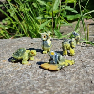 Könnte beinhalten: Vier kleine Tierfiguren aus gesprenkeltem grünem und gelbem Stein. Die Sammlung umfasst eine Schildkröte, eine Eule, einen Elefanten und einen Frosch auf einem Seerosenblatt. Jedes Stück zeigt detaillierte Schnitzereien und kleine orange Augen.