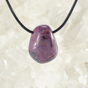 Può includere: Un pendente di rubino lucido a forma di goccia infilato su un cordino nero. La gemma è di un rosso-rosato intenso con sottili variazioni di colore e una superficie liscia e lucida. Il cordino sembra essere in pelle o materiale simile.