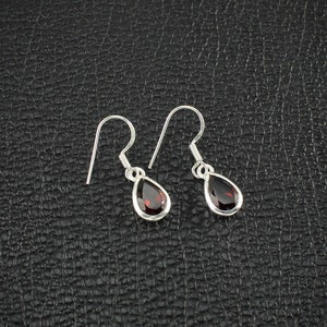 May include: A pair of silver drop earrings with teardrop-shaped, deep red gemstones. The earrings have a simple, elegant design with a hook closure. The gemstones are set in a silver bezel, creating a polished and refined look.