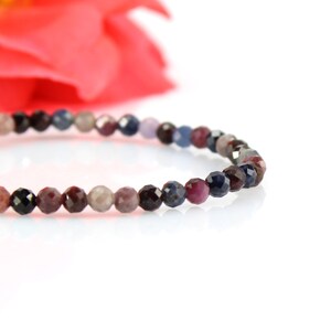 Op de afbeelding: Een delicate armband gemaakt met kleine, gefacetteerde edelsteen kralen in tinten blauw, paars en zwart. De kralen zijn geregen op een doorzichtig elastisch koord.